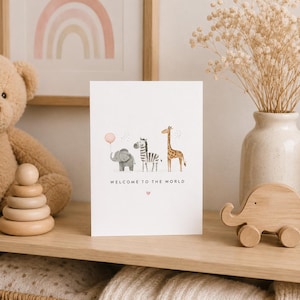 May include: A white greeting card featuring a watercolor illustration of a gray elephant holding a pink balloon, a zebra, and a giraffe. The text "WELCOME TO THE WORLD" is printed in black below the animals, with a small pink heart underneath.