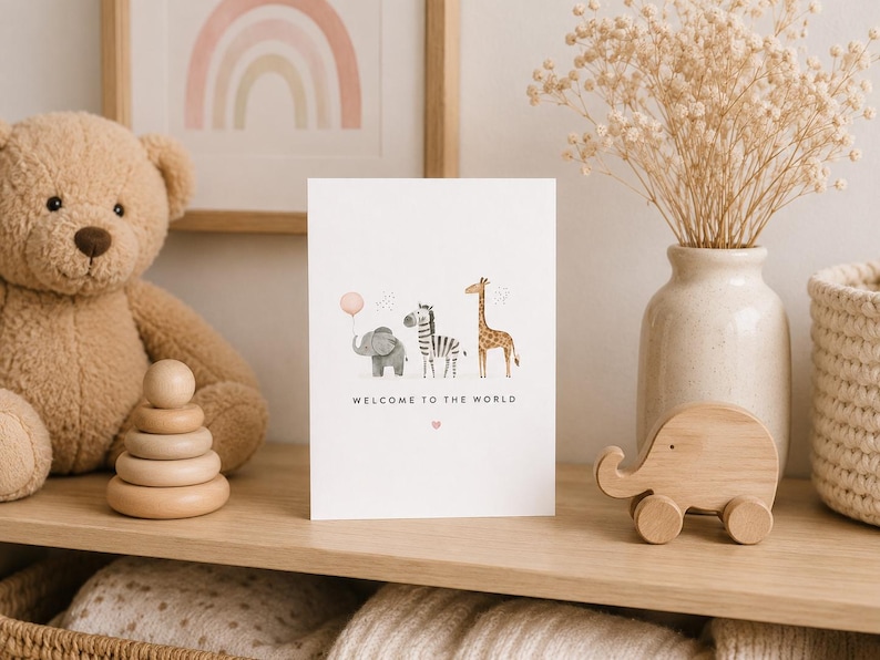 May include: A white greeting card featuring a watercolor illustration of a gray elephant holding a pink balloon, a zebra, and a giraffe. The text "WELCOME TO THE WORLD" is printed in black below the animals, with a small pink heart underneath.