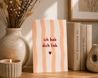 Puede incluir: Una tarjeta de felicitación rectangular con rayas verticales onduladas de color melocotón sobre un fondo crema. El texto "ich hab dich lieb." está impreso en rojo en el centro, seguido de un pequeño icono de corazón rojo.