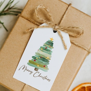 Könnte beinhalten: Ein Weihnachtsgeschenk, verpackt in braunes Papier, mit Bindfaden gebunden und einem weißen Anhänger. Der Anhänger zeigt ein Aquarell-Weihnachtsbaum-Design mit dem Schriftzug "Merry Christmas". Getrocknete Orangenscheiben und Grünzeug sind im Hintergrund.