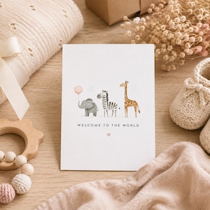 May include: A white greeting card featuring a watercolor illustration of a gray elephant holding a pink balloon, a zebra, and a giraffe. The text "WELCOME TO THE WORLD" is printed in black below the animals, with a small pink heart underneath.