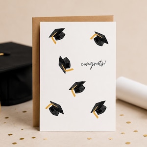 Könnte beinhalten: Eine weiße Grußkarte mit einem Muster aus schwarzen Doktorhüten mit goldenen Quasten und dem handschriftlichen Text congrats! in schwarzer Tinte. Die Karte liegt bei einem braunen Umschlag neben einem schwarzen Doktorhut und einem Diplom.