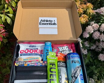 May include: A brown cardboard box with the words "Athlete Essentials" printed on the front. The box is open and contains a variety of athletic snacks and supplements, including energy gels, protein bars, and electrolyte drinks. The box also includes a black fanny pack with a reflective strip.