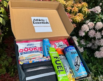 May include: An open cardboard box branded "Athlete Essentials" containing sports nutrition items. Visible products include a cookie, energy gels, a pre-workout drink, and a black waist pack. The box is set against a background of green foliage.