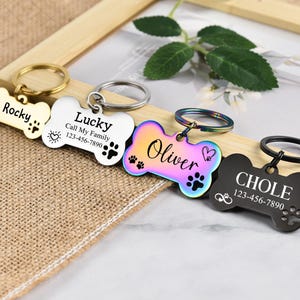 May include: Four dog bone-shaped pet ID tags with engraved names and phone numbers. The tags are in gold, silver, rainbow, and black.
