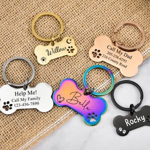 May include: Five different dog bone-shaped pet ID tags with engraved text and paw prints. The tags are in various colors, including silver, gold, black, and iridescent.