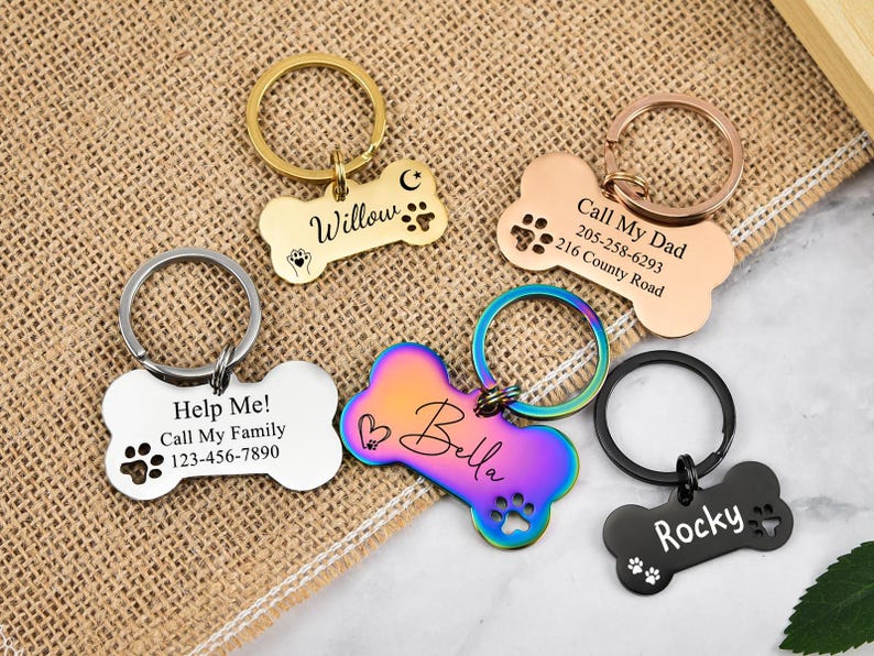 May include: Five different dog bone-shaped pet ID tags with engraved text and paw prints. The tags are in various colors, including silver, gold, black, and iridescent.