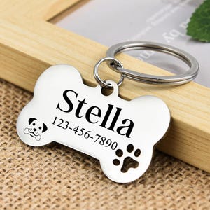 May include: Silver dog tag shaped like a bone with the name Stella and phone number 123-456-7890 engraved on it. The tag has a paw print and a small dog silhouette.