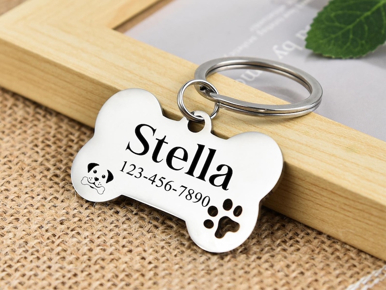 May include: Silver dog tag shaped like a bone with the name Stella and phone number 123-456-7890 engraved on it. The tag has a paw print and a small dog silhouette.