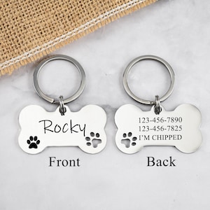 May include: Silver bone-shaped dog tag with a keychain. The front side is engraved with the name "Rocky" and two paw prints. The back side has two phone numbers, the text "I'm chipped", and a paw print.