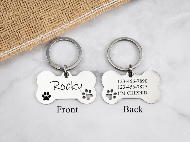 May include: Silver bone-shaped dog tag with a keychain. The front side is engraved with the name "Rocky" and two paw prints. The back side has two phone numbers, the text "I'm chipped", and a paw print.