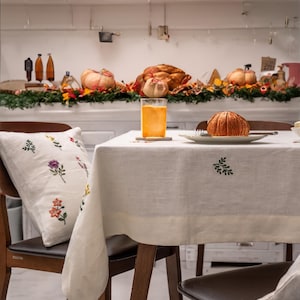 May include: A white tablecloth with embroidered floral designs covers a wooden table with two chairs. A glass of orange juice sits on a coaster on the table, next to a plate with a brown pumpkin-shaped object. The table is set for a fall-themed dinner.
