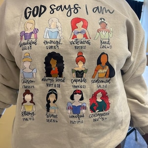 Puede incluir: Una sudadera beige con la frase "GOD says I am" sobre ilustraciones de princesas de dibujos animados. Cada princesa est&aacute; etiquetada con un adjetivo positivo y un vers&iacute;culo b&iacute;blico, como "hermosa" y "elegida".