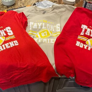 Puede incluir: Tres camisetas con dise&ntilde;os de f&uacute;tbol. Se ven dos camisetas rojas y una sudadera beige. Las camisetas presentan el texto "Taylor's Boyfriend" y "Taylor's" con un gr&aacute;fico de f&uacute;tbol y el n&uacute;mero 87.