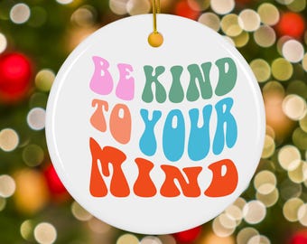 May include: A white ceramic Christmas ornament with the text "BE KIND TO YOUR MIND" in pink, green, orange, and blue retro font. The ornament has a gold string for hanging and is set against a blurred background of Christmas lights.
