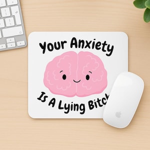 May include: White mousepad with the text "Your Anxiety Is A Lying Bitch" and a pink cartoon brain with a smiling face. A white computer mouse is next to the mousepad.
