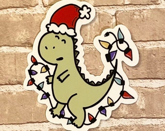 May include: A green dinosaur wearing a red Santa hat is tangled in a string of colorful Christmas lights.