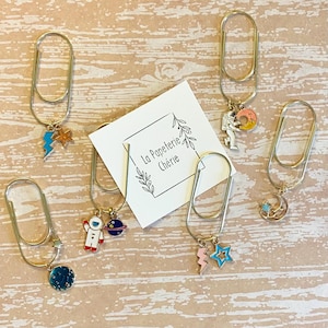 May include: Five gold paper clips with different space-themed charms. The charms include a lightning bolt and star, an astronaut with a planet, a crescent moon with stars, and an astronaut with a moon. The paper clips are on a wooden surface with a white card that says "La Papeterie Chérie".