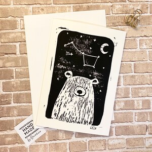May include: Black and white linocut print of a bear looking up at a constellation of stars and a crescent moon. The print is hand-made and signed by the artist.