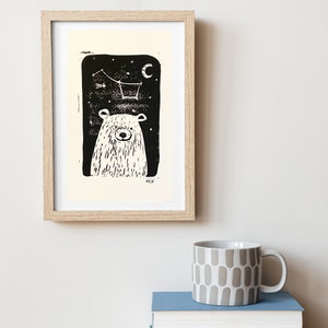 May include: A black and white illustration of a bear looking up at a constellation of stars in the night sky. The bear is framed in a light wood frame.