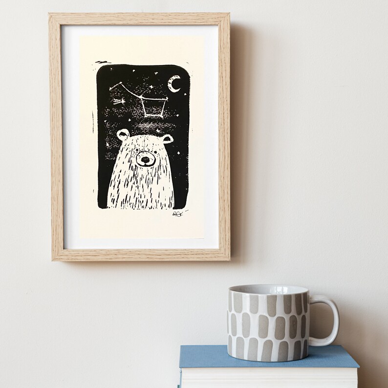 May include: A black and white illustration of a bear looking up at a constellation of stars in the night sky. The bear is framed in a light wood frame.