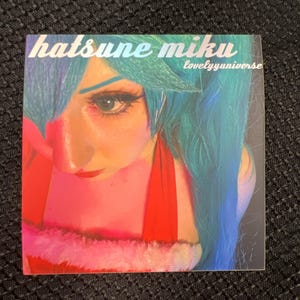 May include: A square image featuring a person with blue hair and makeup, reminiscent of a character. The text "hatsune miku lovelyuniverse" is displayed in a stylized font. The image has a red and pink color scheme.