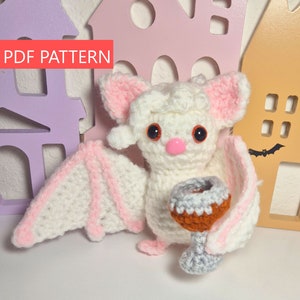 May include: A handmade crochet bat with white and pink wings, ears, and accents, holding a small goblet. The bat has large, dark eyes and a pink nose. The text "PDF PATTERN" is in the upper left corner. The background includes decorative house cutouts.