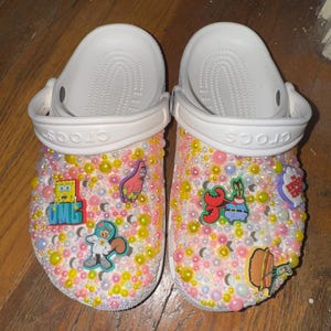 May include: White Crocs shoes customized with colorful beads and cartoon character charms. The charms include SpongeBob SquarePants, Patrick Star, and Mr. Krabs. The shoes are decorated with pink, yellow, and white beads.