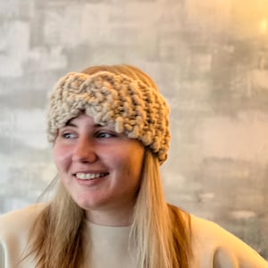 May include: A beige knitted headband with a braided design. The headband is worn by a person with long blonde hair.