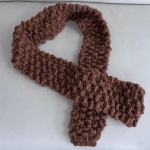 May include: A brown knitted scarf made with a chunky yarn. The scarf is folded in half and tied in a knot.