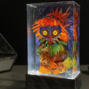 May include: A rectangular, illuminated resin sculpture featuring a colorful character with a mask, orange hair, and green clothing. The sculpture is set on a wooden base and encased in a clear, textured block. The artwork is lit from within, creating a glowing effect.