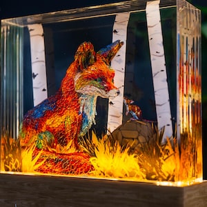 May include: A vibrant fox sculpture within a clear rectangular case, featuring birch tree details. The fox is predominantly red, orange, and yellow, with blue and green accents. The wooden base and warm lighting enhance the scene.