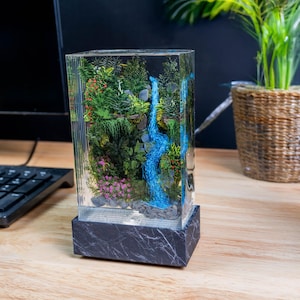 Könnte beinhalten: Rechteckige Kunstharzskulptur mit einer Miniatur-Wasserfallszene mit üppigem Grün, einem blauen Wasserfall und rosa Blüten. Die Skulptur steht auf einem Sockel aus schwarzem und grauem Marmor.
