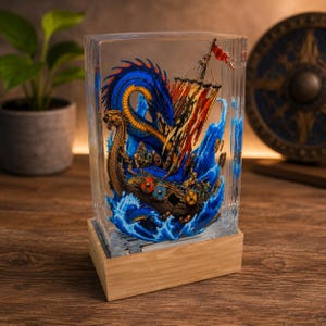 May include: A rectangular clear resin block containing a detailed miniature scene of a wooden Viking ship sailing through blue waves, featuring a large blue sea serpent, colorful sails, and small shields, mounted on a light wood base.