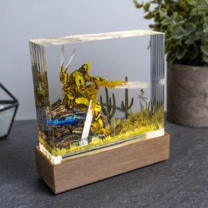 May include: A clear resin sculpture featuring a yellow robot figure amidst a desert scene with miniature cars and cacti. The sculpture is set on a wooden base, creating a unique decorative piece.