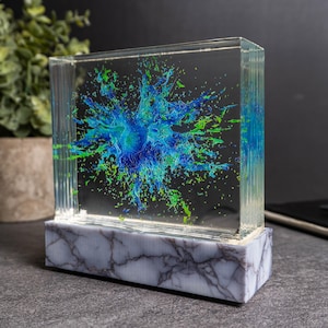 May include: A clear, rectangular art piece with a blue and green splash design. The artwork is encased in a clear block and sits on a marble-patterned base. The piece is a decorative item.
