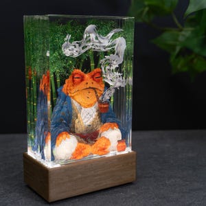 May include: A resin art piece featuring an orange toad in a blue coat, smoking a pipe, set against a bamboo backdrop. The artwork is encased in a clear rectangular block, mounted on a wooden base.