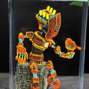 May include: A colorful, wire-framed robot sculpture with a bird perched on its hand. The robot is primarily orange, yellow, and blue, with intricate details. The bird is orange. The sculpture is encased in a clear block.