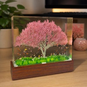 May include: A decorative resin block featuring a miniature pink flowering tree with a light brown trunk, set on a base of green grass and butterflies. The tree is encased in a clear rectangular block, mounted on a dark brown wooden base.
