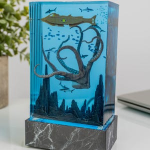 May include: A rectangular resin sculpture featuring an underwater scene. A brown octopus with blue spots and a submarine are suspended in blue resin. The sculpture is set on a marble-patterned base, creating a unique decorative piece.