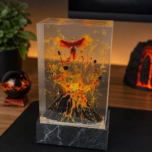 May include: A rectangular, clear resin sculpture featuring a volcano erupting with red and yellow lava, and a phoenix rising above. The sculpture sits on a black and white marble base. A decorative item for a desk or shelf.