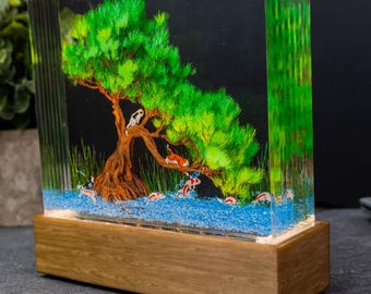 May include: A decorative resin block featuring a miniature landscape with a tree, koi fish, and cats. The tree has green foliage and brown trunk. The base is made of wood. The scene is set against a dark background.