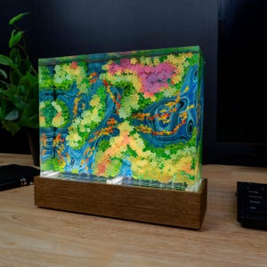 Puede incluir: Una pieza de arte decorativa con un diseño floral colorido encerrado en un bloque rectangular transparente. La obra de arte tiene una base de madera y muestra una vibrante mezcla de colores, incluyendo azul, verde, amarillo, naranja y rosa. La pieza está iluminada, creando un efecto visual llamativo.