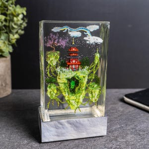 May include: A clear resin block featuring a miniature Japanese pagoda with red and black details, set amidst floating green islands and a blue dragon. The pagoda is surrounded by a pink tree and white clouds. The base is a light gray color.