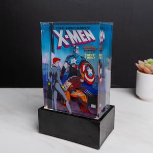 Op de afbeelding: Een helder acrylblok met een vintage stripboekcover van "The Uncanny X-Men". De cover toont Captain America, Black Widow en Wolverine. Het blok staat op een zwarte rechthoekige basis.