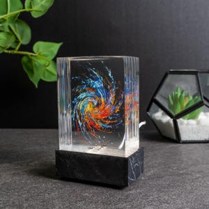 Pode incluir: Lâmpada retangular transparente com um design de espiral abstrato colorido em azul, laranja e amarelo. A lâmpada está sobre uma base preta com efeito mármore. A lâmpada está ligada e acesa.