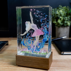 Könnte beinhalten: Rechteckiger dekorativer Harzblock mit einer Ballerina in einem rosa Tutu, umgeben von blauen und rosa floralen Akzenten. Das Kunstwerk befindet sich vor einem schwarzen Hintergrund und steht auf einem Holzsockel. Der Block ist transparent.
