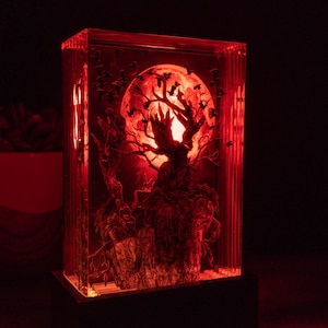 May include: A red-lit, rectangular art piece featuring a tree silhouette against a full moon. The tree's branches reach towards the moon, with several birds in flight. The artwork is encased in a clear, rectangular block.