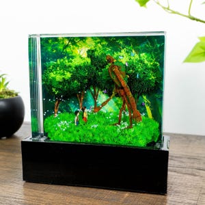 Könnte beinhalten: Ein Diorama mit einer Waldszene und einem großen Holzroboter. Zwei Kinder stehen in einer grasbewachsenen Gegend, Schmetterlinge fliegen um den Roboter. Das Diorama befindet sich in einem transparenten, rechteckigen Rahmen und steht auf einem schwarzen Sockel.