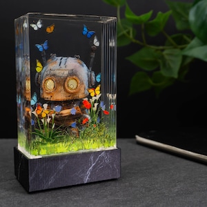 Könnte beinhalten: Ein klarer Kunstharzblock mit einer detaillierten Roboterfigur, umgeben von bunten Schmetterlingen und Blumen. Der Roboter hat ein verwittertes, metallisches Aussehen mit großen, kreisförmigen Augen. Der Sockel ist ein dunkler Block mit Marmormuster.